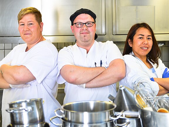 Drei Küchenmitarbeitende in weißer Arbeitskleidung stehen nebeneinander in einer Großküche, die Arme selbstbewusst verschränkt. Die mittlere Person trägt Mütze und Brille. Im Vordergrund stehen glänzende Kochtöpfe und Küchenutensilien. Die Szene vermittelt Teamarbeit und Professionalität.