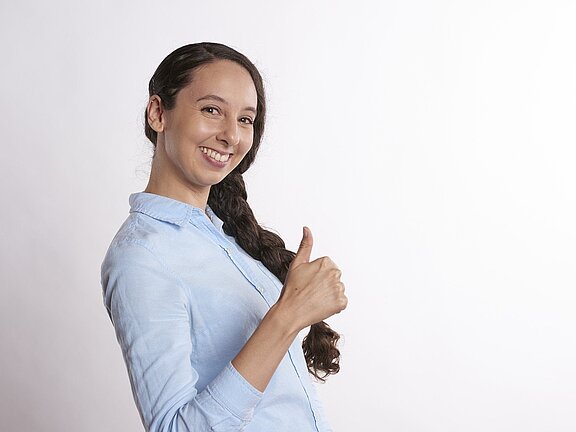Eine lächelnde Frau zeigt mit ihrer rechten Hand ein Daumenhoch. Sie trägt ein hellblaues Hemd und hat lange, dunkelbraune Haare, die zu einem Zopf geflochten sind. Der Hintergrund ist hell und neutral.