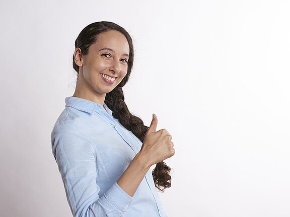 Eine lächelnde Frau zeigt mit ihrer rechten Hand ein Daumenhoch. Sie trägt ein hellblaues Hemd und hat lange, dunkelbraune Haare, die zu einem Zopf geflochten sind. Der Hintergrund ist hell und neutral.
