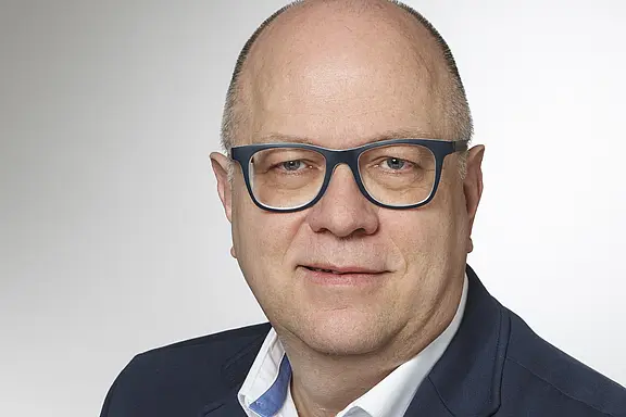 Porträtfoto: Mann mittleren Alters mit Glatze und dunkelblauer Brille, trägt dunkelblaues Sakko und weißes Hemd, schaut in die Kamera und lächelt leicht; neutraler, heller Hintergrund.