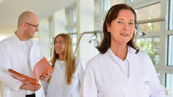 Drei medizinische Fachkräfte in weißen Kitteln im hellen Krankenhausflur; eine Frau steht vorn und lächelt, im Hintergrund unterhalten sich zwei Kolleg*innen mit Akten. Der Fokus liegt auf Professionalität und Teamarbeit.