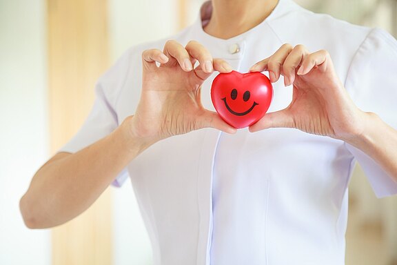 A healthcare worker in a white uniform holds a small red heart with a smiling face between both hands, symbolizing caring and positivity.
