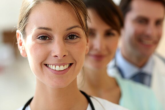 A smiling female healthcare professional appears in the foreground, wearing a stethoscope. Two other healthcare workers are visible in the background, slightly out of focus, creating a sense of teamwork and support in a medical environment.