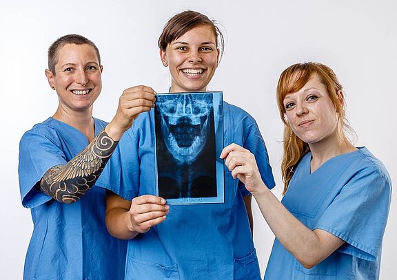 Drei Frauen in blauen Ärzteschürzen stehen zusammen und lächeln. Eine von ihnen hält ein Röntgenbild eines Kiefers in der Hand. Die anderen beiden schauen interessiert auf das Bild, während sie ihre Unterstützung zeigen. Die Atmosphäre ist freundlich und professionell.