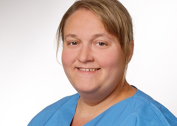 Kopf-und-Schulter-Porträt einer lächelnden Person in blauer medizinischer Kleidung vor neutralem Hintergrund.