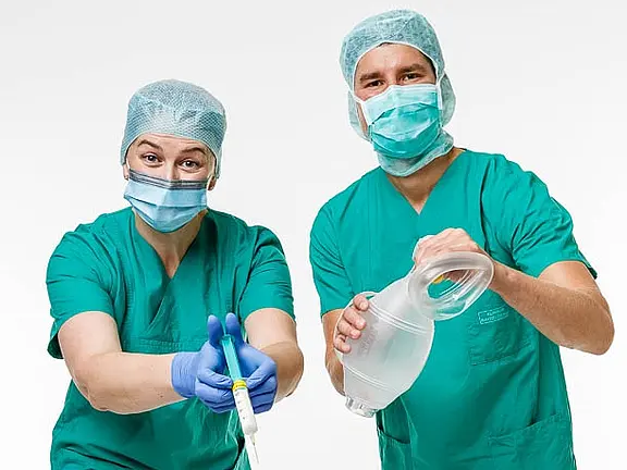 Zwei medizinische Fachkräfte in grünen Scrubs und OP-Hauben stehen zusammen. Die Frau hält eine Spritze und zeigt darauf, während der Mann einen medizinischen Behälter in der Hand hält. Beide tragen Mund-Nasen-Schutz und schauen freundlich in die Kamera.