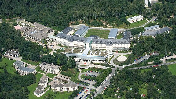 Aerial view of a large, U-shaped institutional complex with slate roofs and a modern glass central section, set within dense greenery. Curved driveways and parking lots line the front, with several smaller buildings nearby.