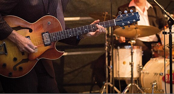 Nahaufnahme eines Gitarristen, der eine halbakustische E-Gitarre spielt; im Hintergrund sitzt ein Schlagzeuger am Drumset. Konzertszene mit Bühnenbeleuchtung, der Fokus liegt auf den Händen und Saiten des Gitarristen.