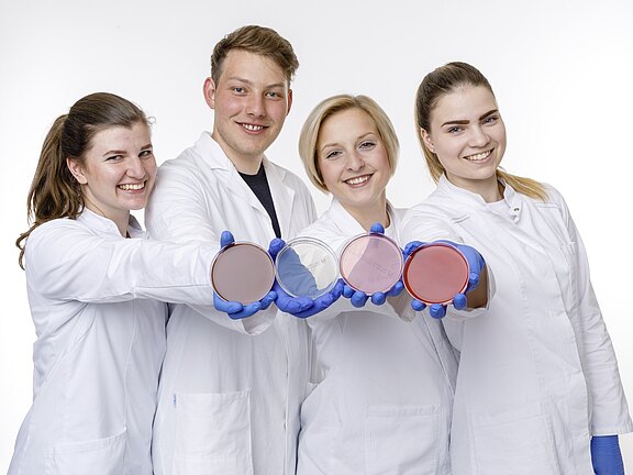 Vier junge Personen im Labor tragen weiße Kittel und blaue Handschuhe. Sie halten vier Petrischalen mit unterschiedlich gefärbten Agarplatten dem Betrachter entgegen und lächeln.