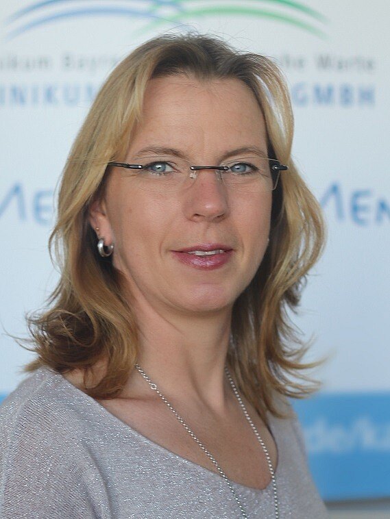 Porträt einer Frau mit blonden, lockigen Haaren, die eine helle Bluse trägt. Sie hat eine freundliche Miene und trägt eine Brille mit dünnem Rahmen. Der Hintergrund ist unscharf, zeigt aber den Schriftzug einer Institution.