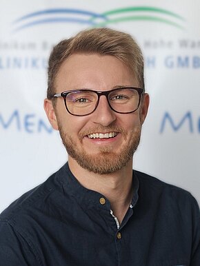 Jens Jungwirth Porträtfoto eines lächelnden jungen Mannes mit Brille und kurzem Bart. Er trägt ein dunkelblaues Hemd und blickt direkt in die Kamera vor neutralem, unscharfem Hintergrund.
