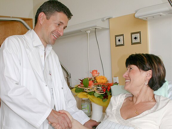 A doctor in a white coat smiles while shaking hands with a female patient in a hospital room. The patient appears cheerful and engaged in conversation. A colorful bouquet of flowers is visible in the background, adding a warm atmosphere to the scene.