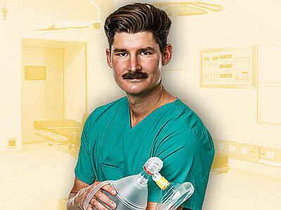Ein junger Mann mit einem Schnurrbart trägt grüne OP-Kleidung und hält ein Beatmungsgerät in den Händen. Im Hintergrund sind verschwommene Elemente eines Krankenhausraums erkennbar, was die medizinische Umgebung unterstreicht.