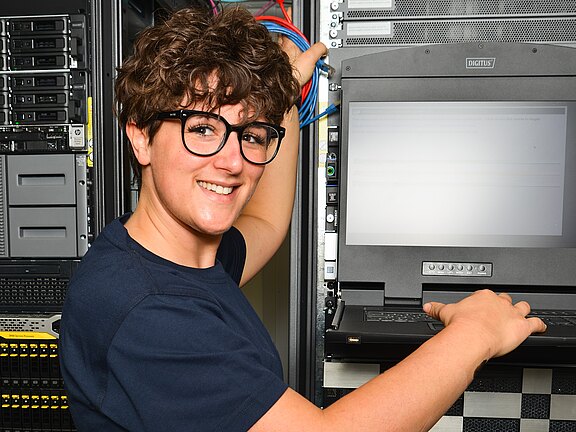 Eine Person im Serverraum wartet einen Serverschrank: Die Person steckt Netzwerkkabel ein und bedient eine ausklappbare Rack-Konsole mit Bildschirm und Tastatur. Fokus: IT-Wartung und Administration an Hardware im Rechenzentrum.