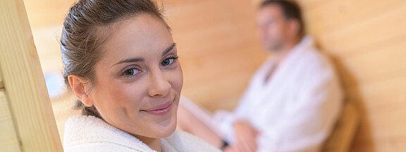 Smiling woman in a white bathrobe relaxing in a wooden sauna; a man in a robe sits blurred in the background.