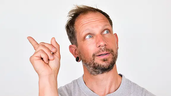 Ein Mann vor hellem Hintergrund blickt nach oben links und zeigt mit erhobenem Zeigefinger dorthin, als wolle er auf etwas hinweisen oder eine Idee haben.