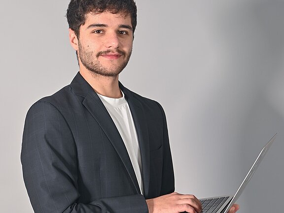Ein junger Mann mit schwarzem Blazer und weißem T-Shirt steht mit einem Laptop in der Hand. Er hat einen freundlichen Ausdruck und schaut in die Kamera. Der Hintergrund ist hell und neutral.