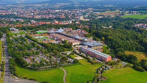 Aerial view of a large campus-like complex with several modern low-rise buildings, parking lots, and tree-lined paths, bordered by parkland and forest. A city spreads across the background, with fields and hills beyond.