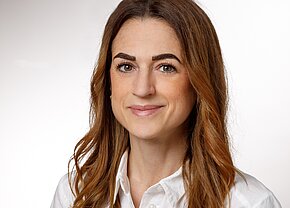 Anna-Lena Gevis Nahaufnahme einer lächelnden Frau mit langen, lockigen braunen Haaren. Sie trägt ein weißes Hemd und hat eine freundliche Ausstrahlung. Der Hintergrund ist hell und neutral.