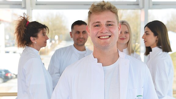 Eine Gruppe von vier Personen in Weiß, wahrscheinlich medizinisches Personal. Ein junger Mann steht lächelnd im Vordergrund, während die anderen im Hintergrund stehen und sich unterhalten. Der Raum hat große Fenster, durch die Tageslicht strömt.