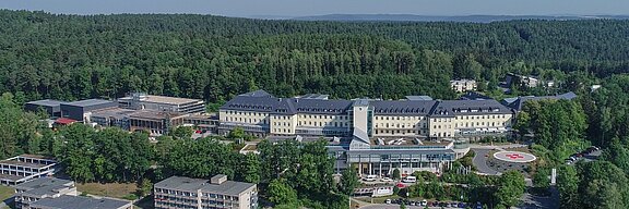 Luftaufnahme eines großen Gebäudekomplexes, umgeben von dichtem Wald. Im Vordergrund befindet sich ein zentraler Eingangsbereich mit einer roten Kreuzsymbolik. Die Architektur ist modern und farblich hell, eingebettet in eine ruhige, grüne Umgebung.