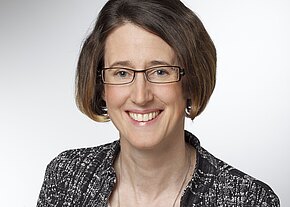 Porträtfoto: Eine lächelnde Frau mit Brille und kurzem braunem Haar, in einem gemusterten Blazer und mit Kette. Neutraler Hintergrund, Fokus auf das Gesicht in professioneller Pose.