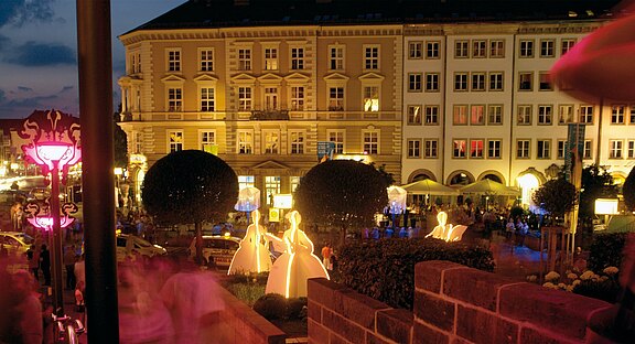 Eine beleuchtete Platzszene bei Nacht, mit eleganten, hellen Silhouetten von Personen in weißen Kleidern. Umgeben von gepflegten Büschen und Gebäude im Hintergrund, das die Atmosphäre eines belebten, festlichen Abends widerspiegelt.