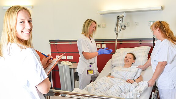 Krankenhauszimmer: Ein Patient liegt lächelnd im Bett. Drei Pflegekräfte in weißer Kleidung betreuen ihn – eine notiert etwas, eine zieht Handschuhe an, eine richtet Bett oder Decke. Die Szene vermittelt medizinische Versorgung und freundliche Atmosphäre.