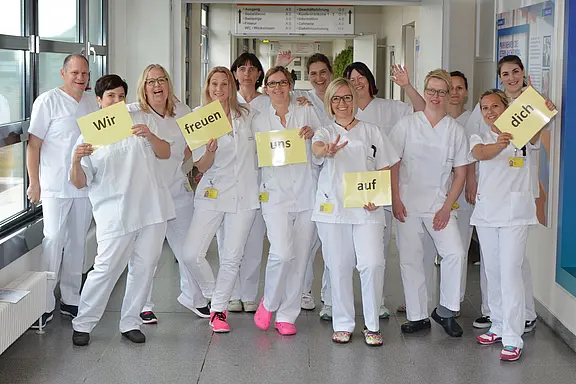 Eine Gruppe Pflegekräfte in weißer Kleidung steht lachend in einem Krankenhausflur und hält Schilder mit der Botschaft: „Wir freuen uns auf dich.“