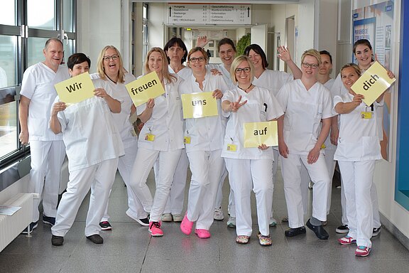 Eine Gruppe Pflegekräfte in weißer Kleidung steht lachend in einem Krankenhausflur und hält Schilder mit der Botschaft: „Wir freuen uns auf dich.“