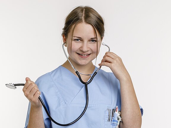 Porträt einer lächelnden medizinischen Fachkraft in hellblauer Kleidung. Sie hält ein Stethoskop, hat die Ohrbügel eingesetzt und präsentiert das Instrument in der Hand. Neutraler Hintergrund.