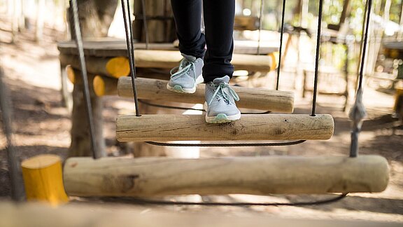 Nahaufnahme: Füße in Turnschuhen treten über schwebende Holzstämme einer Seilbrücke im Kletterpark; die Person balanciert vorsichtig.