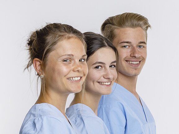 Drei freundlich lächelnde Personen in hellblauen Pflegeuniformen stehen nebeneinander im Halbprofil vor weißem Hintergrund.