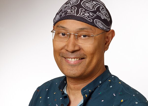 A smiling man is wearing a dark patterned bandana and glasses. He has short hair and is dressed in a teal shirt with colorful patterns. The background is a light gradient that enhances his cheerful expression.