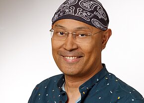 Aaron Warnke-Marinay A smiling man is wearing a dark patterned bandana and glasses. He has short hair and is dressed in a teal shirt with colorful patterns. The background is a light gradient that enhances his cheerful expression.