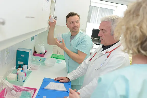 Two healthcare professionals in scrubs discuss a clear container held by one of them. They are in a clinical setting, studying notes on a blue folder, while a third person in scrubs observes them.