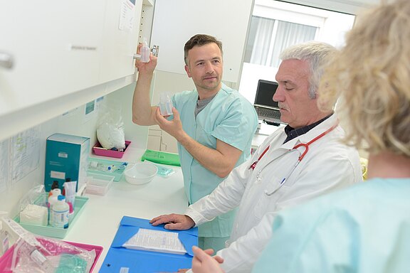 Two healthcare professionals in scrubs discuss a clear container held by one of them. They are in a clinical setting, studying notes on a blue folder, while a third person in scrubs observes them.