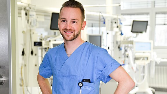 Ein junger Mann in blauem Arztkittel steht lächelnd in einem Krankenhausumfeld. Er hat eine kurze Frisur und einen Bart. In seiner Kitteltasche befinden sich ein Schlüsselanhänger und eine Wasserflasche. Im Hintergrund sind medizinische Geräte zu sehen.