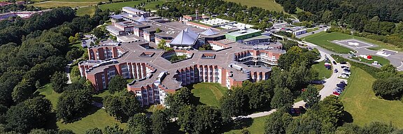 Luftaufnahme eines modernen Gebäudes, das von einer grünen Landschaft umgeben ist. Das Gebäude hat eine einzigartige, sternförmige Architektur mit mehreren Eingängen und ist mit Bäumen und Rasenflächen umgeben. Ein Parkplatz ist in der Nähe sichtbar.