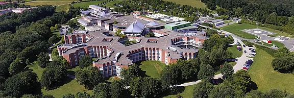 Luftaufnahme eines modernen Gebäudes, das von einer grünen Landschaft umgeben ist. Das Gebäude hat eine einzigartige, sternförmige Architektur mit mehreren Eingängen und ist mit Bäumen und Rasenflächen umgeben. Ein Parkplatz ist in der Nähe sichtbar.