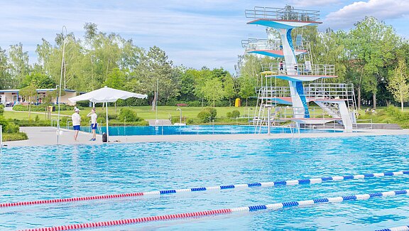 Großes Freibad mit Schwimmbahnen im Vordergrund. Rechts steht ein mehrstöckiger Sprungturm über dem Wasser. Sommerliche Szene mit wenigen Badegästen; Bäume und Wiese im Hintergrund.